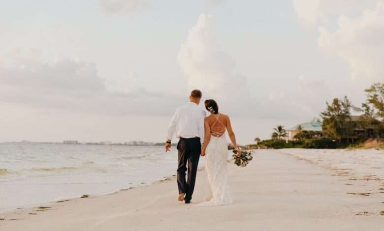 A bride and groom walk away on the beach as they hold hands. They're leaning in toward one another.