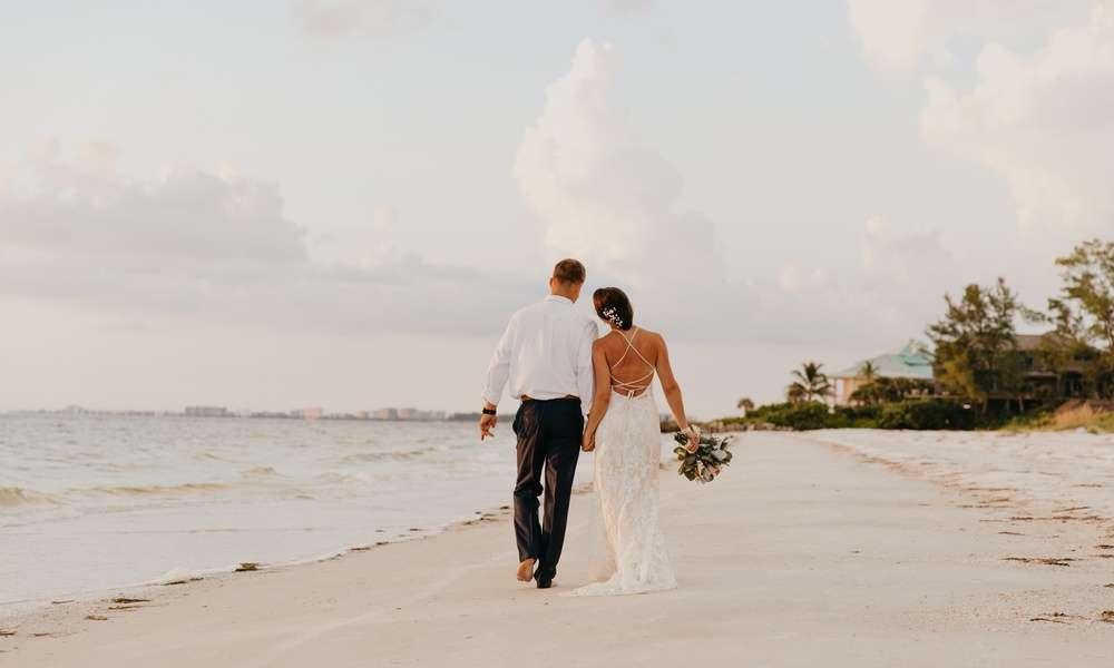 A bride and groom walk away on the beach as they hold hands. They're leaning in toward one another.