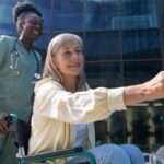 A nurse wearing green scrubs pushes a patient in a wheelchair up to a recycling bin for her to throw out her coffee cup.