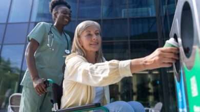 A nurse wearing green scrubs pushes a patient in a wheelchair up to a recycling bin for her to throw out her coffee cup.