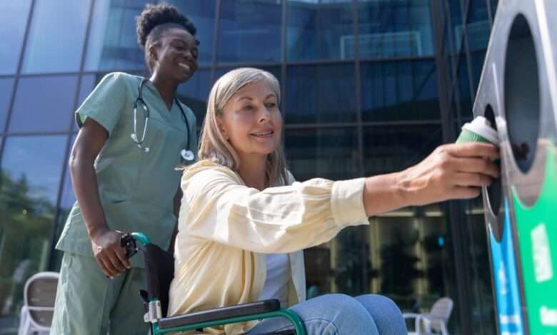 A nurse wearing green scrubs pushes a patient in a wheelchair up to a recycling bin for her to throw out her coffee cup.