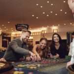 A dealer and smiling players are gathered around a roulette table. Some players move their stacks of chips on the table.