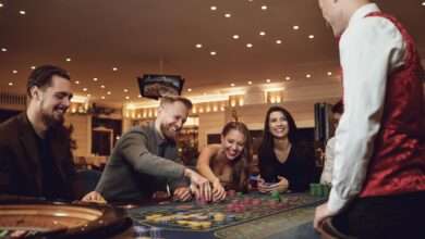 A dealer and smiling players are gathered around a roulette table. Some players move their stacks of chips on the table.