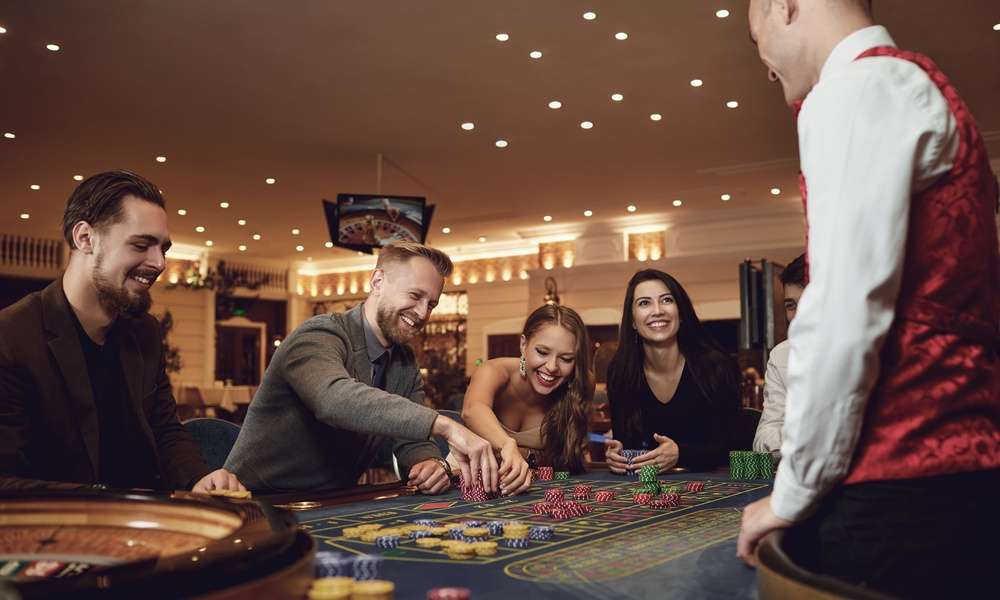 A dealer and smiling players are gathered around a roulette table. Some players move their stacks of chips on the table.
