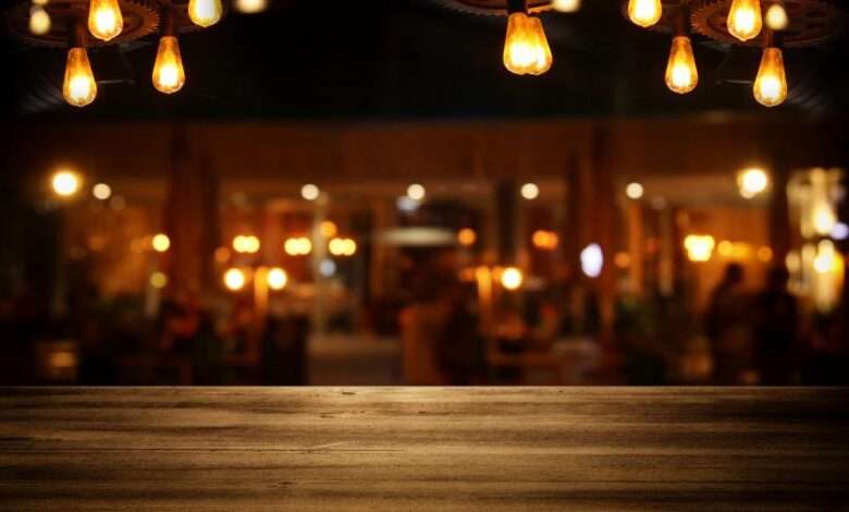 A close-up of a bar top with a blurred view of the bar's interior. There are Edison bulbs hanging from light fixtures.