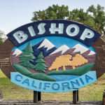 A sign featuring mountains, a river, and trees, with the words Bishop California. The sign sits in a natural area.
