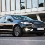 A beautiful black sedan car is parked in front of a building. It is shiny, free of dings and dents, and looks like new.