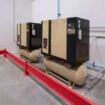 Two beige and black air compressor units and a yellow bucket in a building with white walls and cement floors.