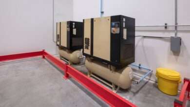 Two beige and black air compressor units and a yellow bucket in a building with white walls and cement floors.