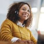 A happy young woman working in a yellow sweater and headset, smiling while working on a company call.