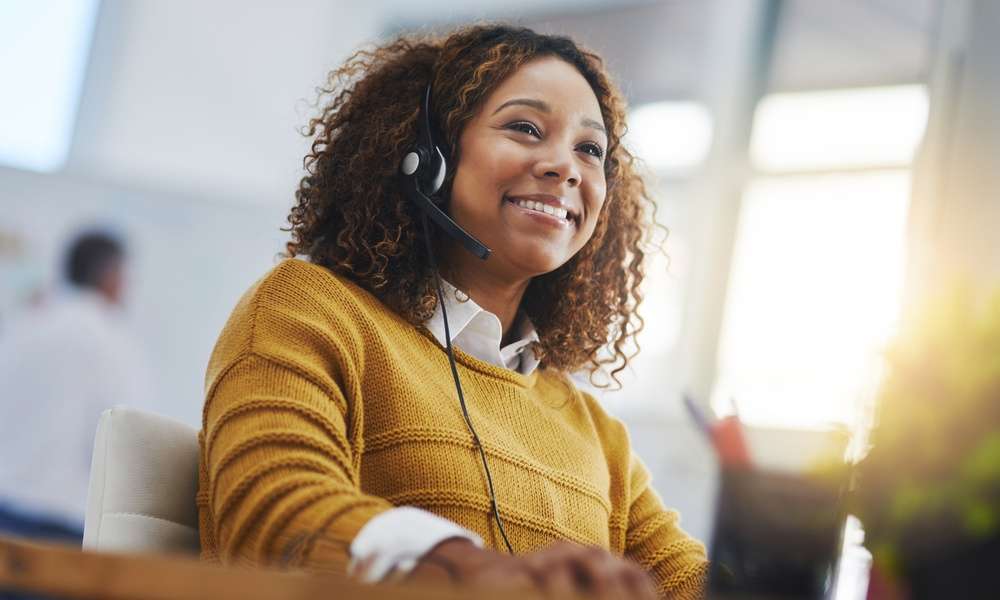 A happy young woman working in a yellow sweater and headset, smiling while working on a company call.