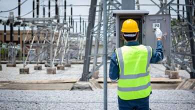 An engineer is inspecting electrical equipment at a high-voltage substation, wearing safety gear and a yellow helmet.