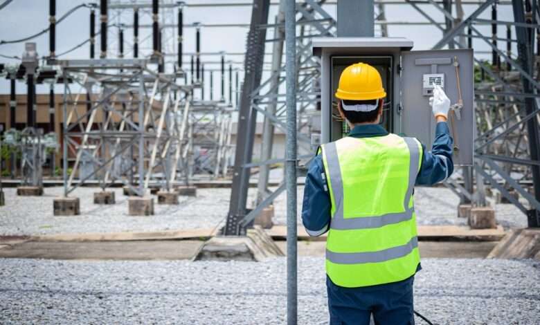 An engineer is inspecting electrical equipment at a high-voltage substation, wearing safety gear and a yellow helmet.