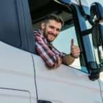 A truck driver, wearing a red and blue flannel, is smiling as he gives the camera a thumbs-up outside the window.