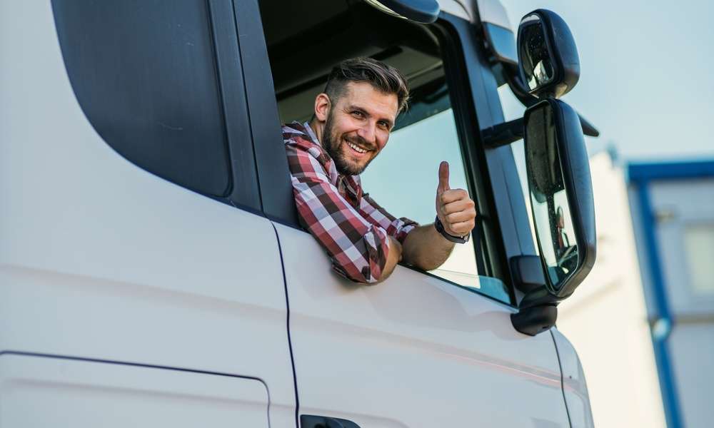 A truck driver, wearing a red and blue flannel, is smiling as he gives the camera a thumbs-up outside the window.