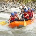 A group of men and women in red life jackets, black pants, and blue helmets rafting through a turbulent river.