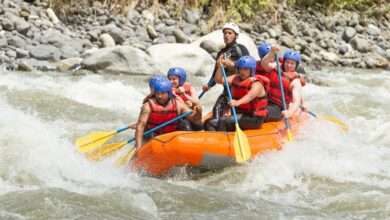 A group of men and women in red life jackets, black pants, and blue helmets rafting through a turbulent river.