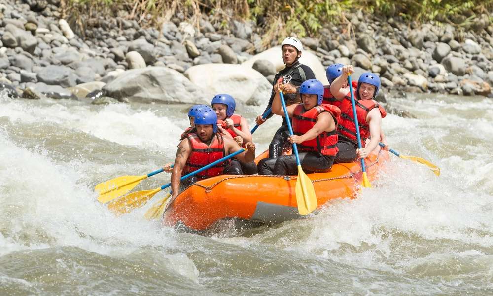 A group of men and women in red life jackets, black pants, and blue helmets rafting through a turbulent river.