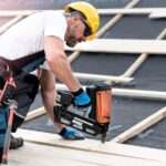 A man in a hard hat, safety glasses, gloves, and overalls uses a large nail gun to fasten pieces of wood.