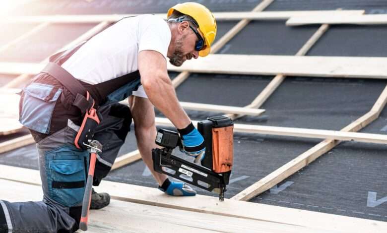 A man in a hard hat, safety glasses, gloves, and overalls uses a large nail gun to fasten pieces of wood.