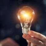 Close-up of the hand of a man wearing a black shirt holding a lightbulb, releasing light with three fingers.