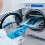 A female laboratory worker wearing blue gloves and a white lab coat puts instruments into an autoclave for sanitation.