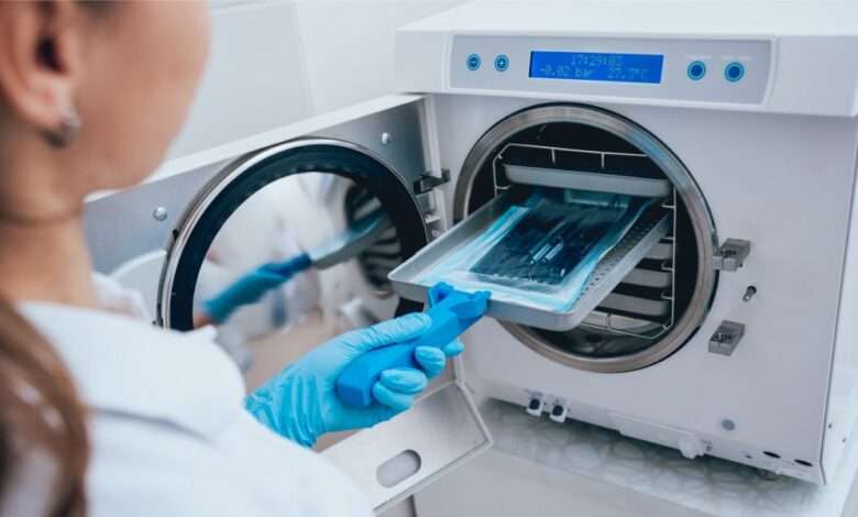 A female laboratory worker wearing blue gloves and a white lab coat puts instruments into an autoclave for sanitation.