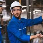 Two men wearing blue industrial work suits and hard hats inspect a piece of equipment in a facility.