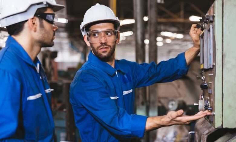 Two men wearing blue industrial work suits and hard hats inspect a piece of equipment in a facility.