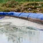 A close-up of a chemical spill on the ground near a grassy area. There is a long, inflated blue barrier stopping the spill.