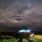 A vehicle parked at a campsite. Stars are visible in the night sky and a light is emitting from inside the vehicle.