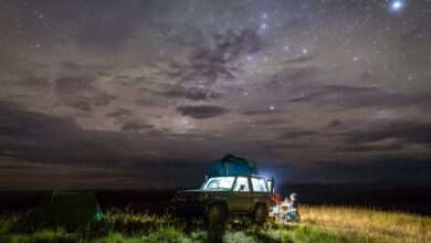 A vehicle parked at a campsite. Stars are visible in the night sky and a light is emitting from inside the vehicle.