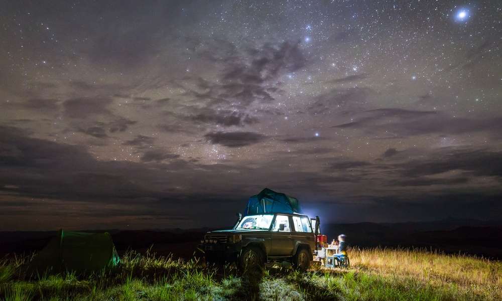 A vehicle parked at a campsite. Stars are visible in the night sky and a light is emitting from inside the vehicle.