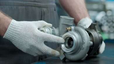 A person wearing knit white gloves holds a vehicle’s turbocharger on a workshop bench before installation.