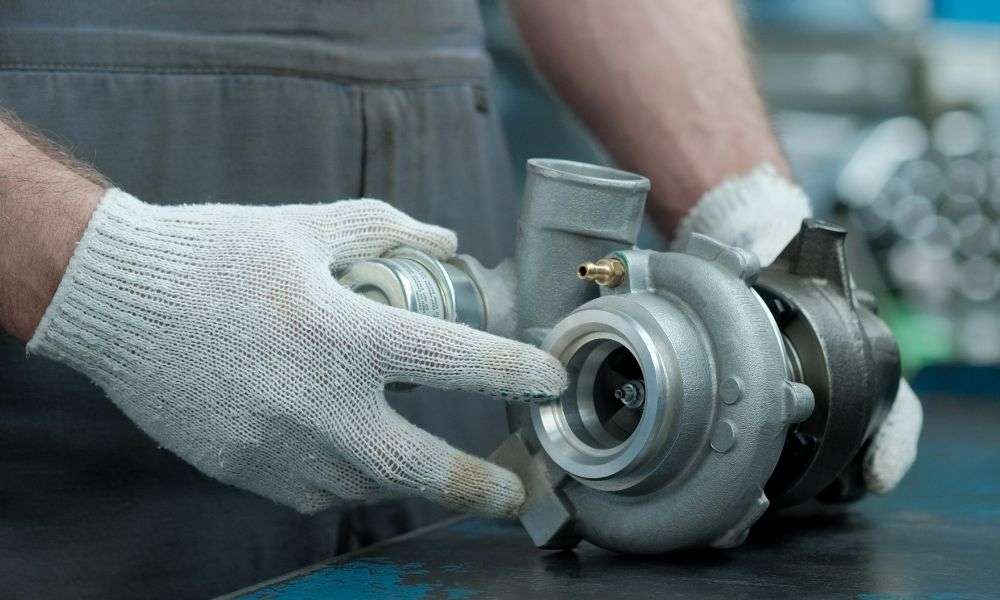 A person wearing knit white gloves holds a vehicle’s turbocharger on a workshop bench before installation.
