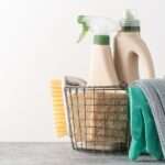 A close-up of brushes, sponges, rubber gloves, and natural cleaning products in a silver metal basket.