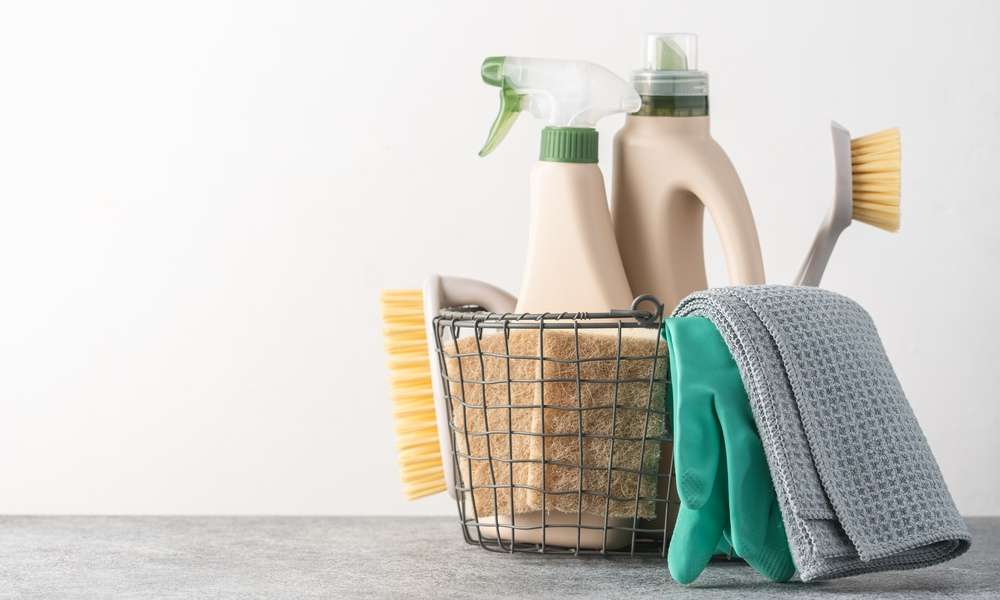 A close-up of brushes, sponges, rubber gloves, and natural cleaning products in a silver metal basket.