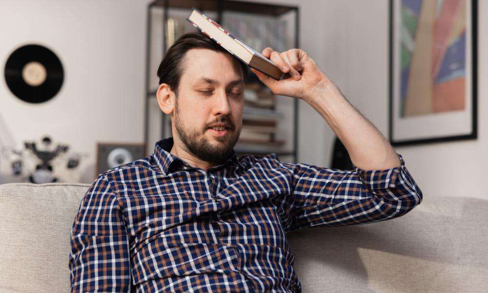 A close-up of a bearded man sits on the couch and hits his head with a dictionary, hoping that he will finally remember the definition.