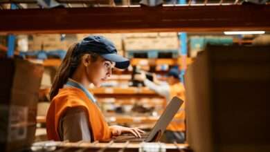 From inside a shelving unit, a warehouse worker is on a laptop. Two boxes on the shelf frame her as she works.