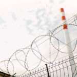 A barbed wire fence at an industrial site. A white smokestack with three red horizontal stripes towers in the distance.