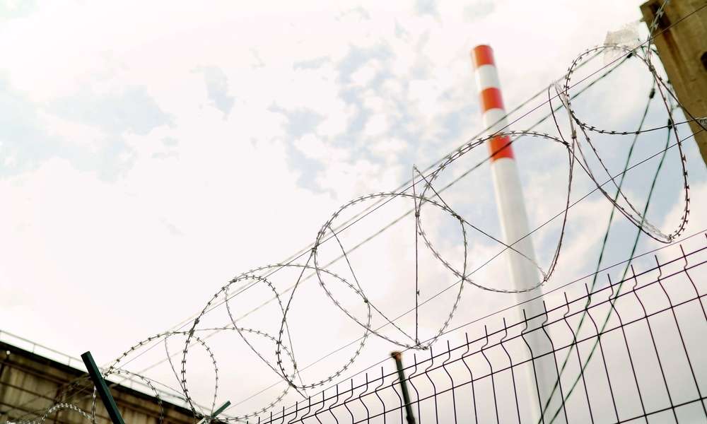 A barbed wire fence at an industrial site. A white smokestack with three red horizontal stripes towers in the distance.