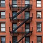 A close-up of an old red-brown brick apartment building in New York City. The fire escape staircase is black.