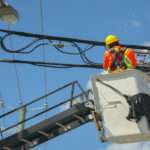A utility worker on a lift while looking at a utility pole's wires. He is wearing a safety vest and a hard hat.