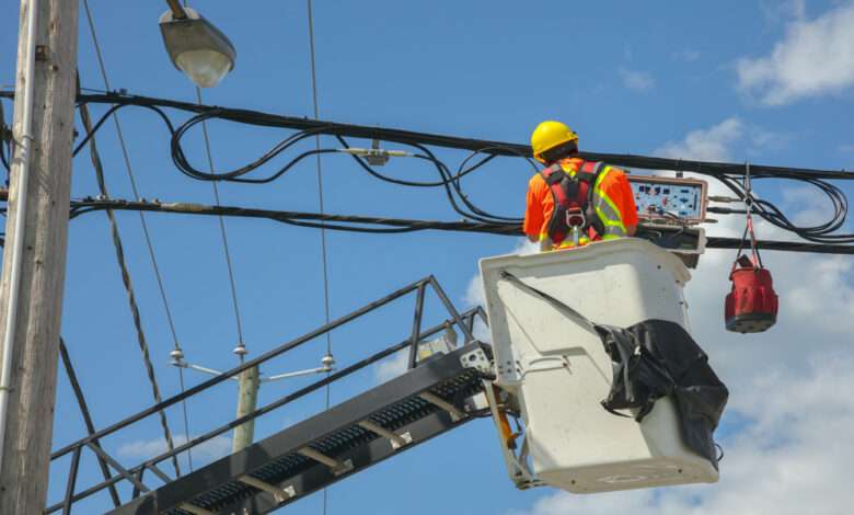 A utility worker on a lift while looking at a utility pole's wires. He is wearing a safety vest and a hard hat.
