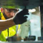 A man wearing safety goggles, a yellow safety vest, and a work glove on his hand works on a machine in a facility.