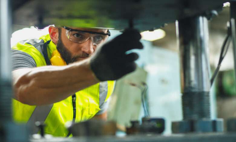 A man wearing safety goggles, a yellow safety vest, and a work glove on his hand works on a machine in a facility.