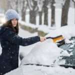 A woman using a brush to wipe snow off her car windshield. There is snow covering the ground around her.