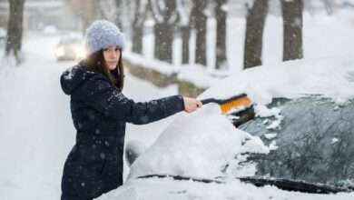 A woman using a brush to wipe snow off her car windshield. There is snow covering the ground around her.