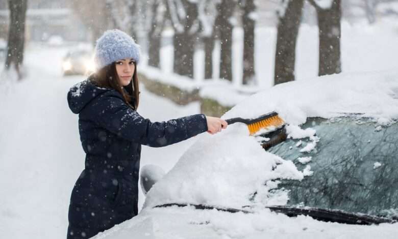 A woman using a brush to wipe snow off her car windshield. There is snow covering the ground around her.