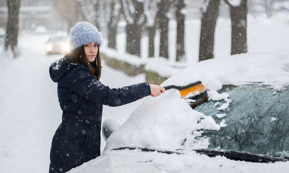 A woman using a brush to wipe snow off her car windshield. There is snow covering the ground around her.
